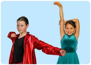 Two young girls pose energetically with raised arms wearing a red shiny jacket and a teal dress against a light blue background