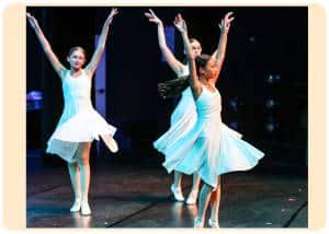 Four young female ballet dancers in white costumes perform on a dark stage arms raised in a graceful choreographed pose